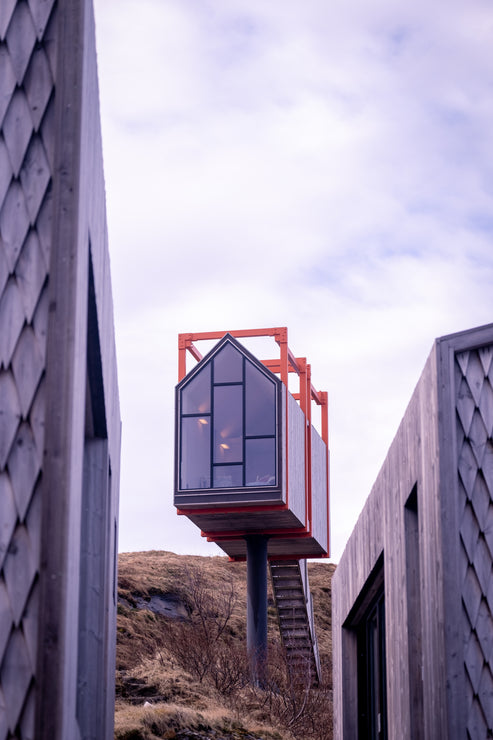 Small geometric house on stilts surrounded by tall buildings under a cloudy sky.